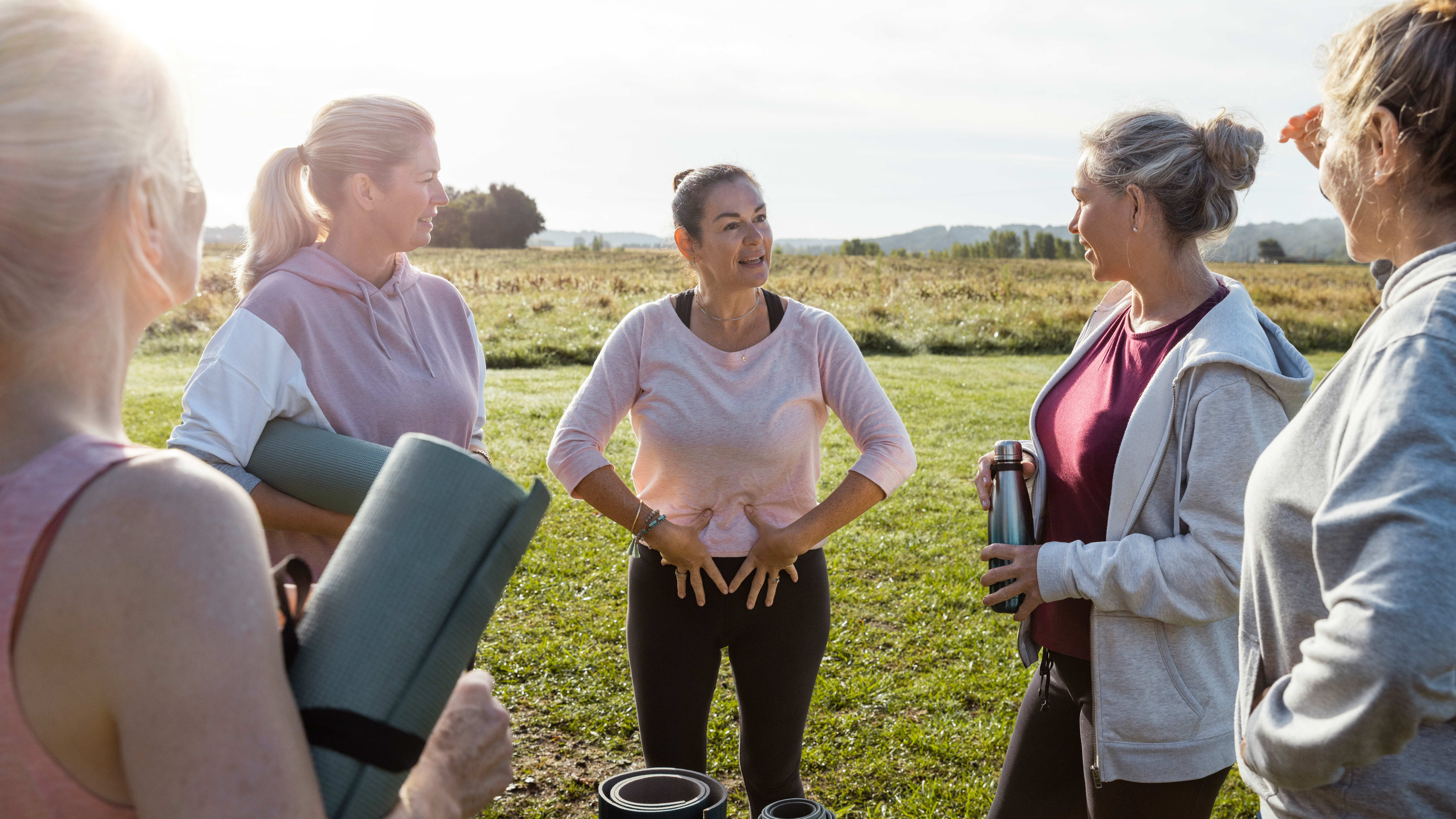A group of women in a grassy field stand talking and holding yoga mats.