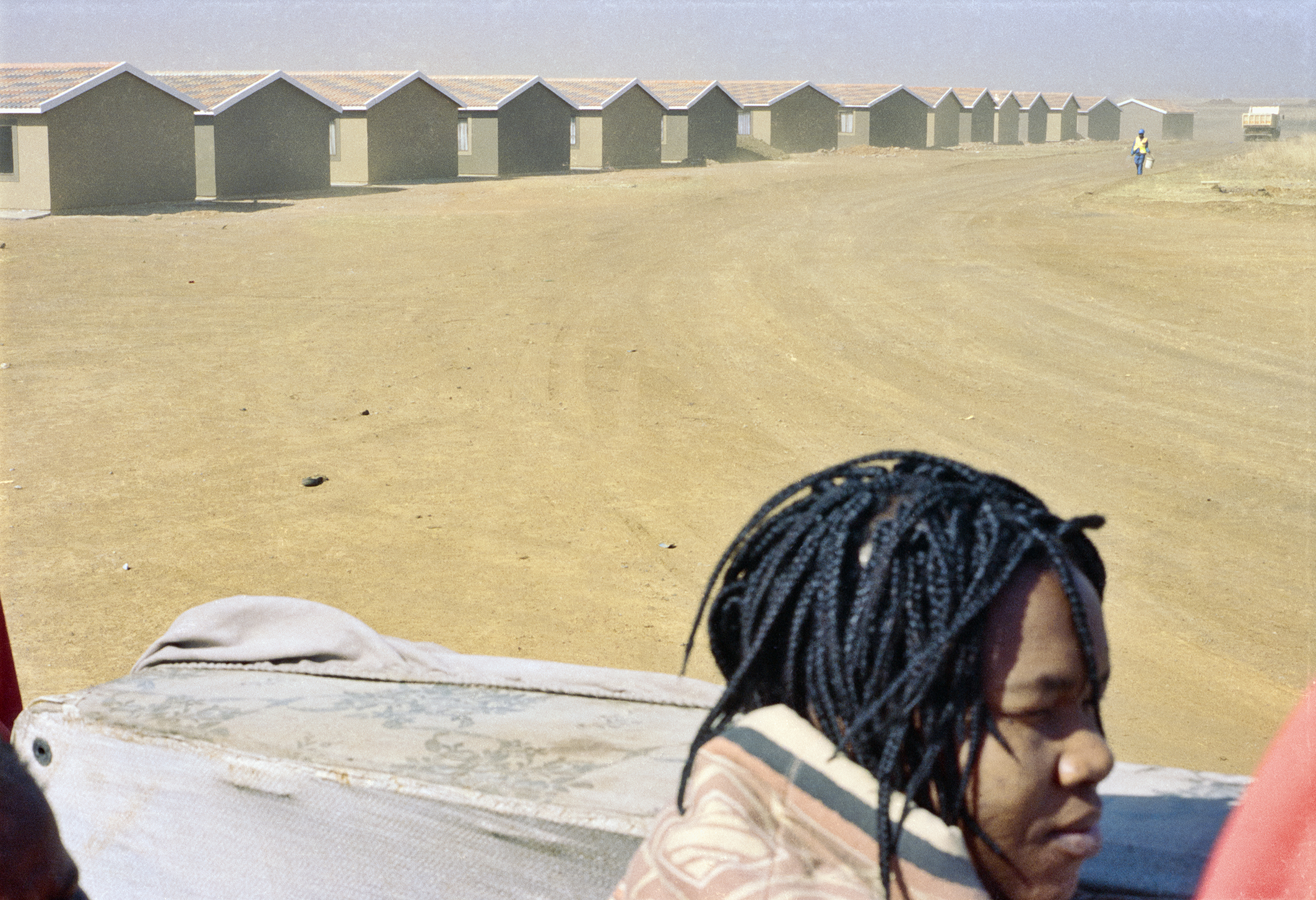 Color photograph of a row of houses on a sandy road. A girl's head can be seen in the corner of the photograph