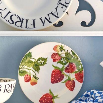 A classic strawberry print Emma Bridgewater plate with a white plate with black Emma Bridgewater font on, against a light blue wall.