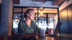 A woman in a smart green top sitting in a meeting room in an office in front of her laptop, which is on a table, laughing while on a video call