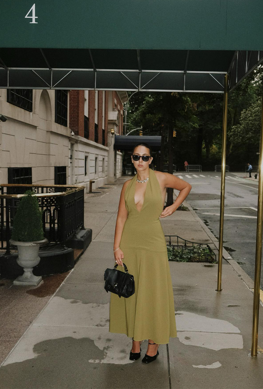 halter dress trend on a woman wearing a green midi dress styled with black ankle strap pumps, a silver conch-shaped necklace, a black handbag, and black sunglasses
