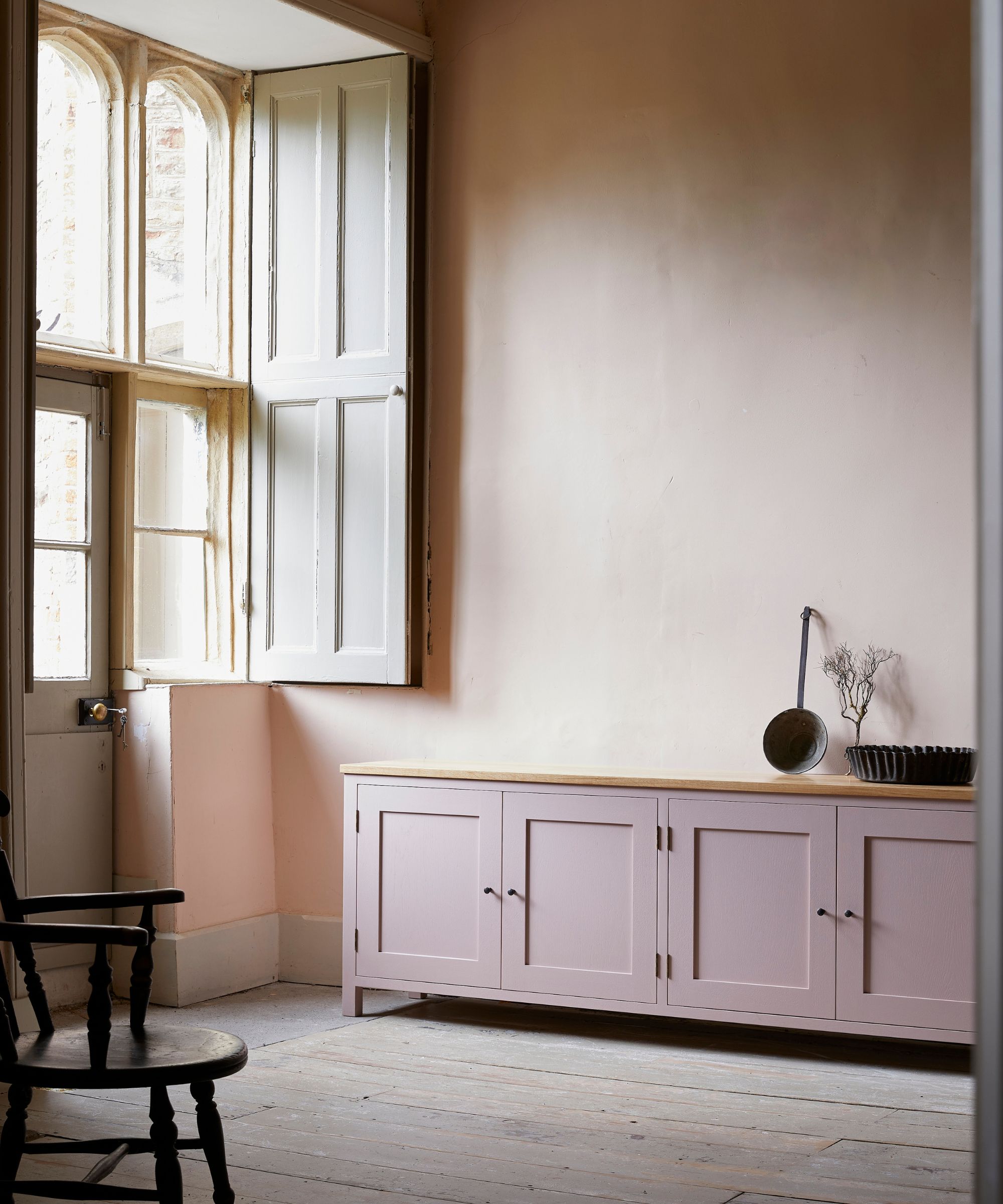 A large hallway with plaster walls, large windows with beige wooden shutters, and exposed wooden floorboards, a pink painted shaker console and a dark wood shaker chair in the corner.