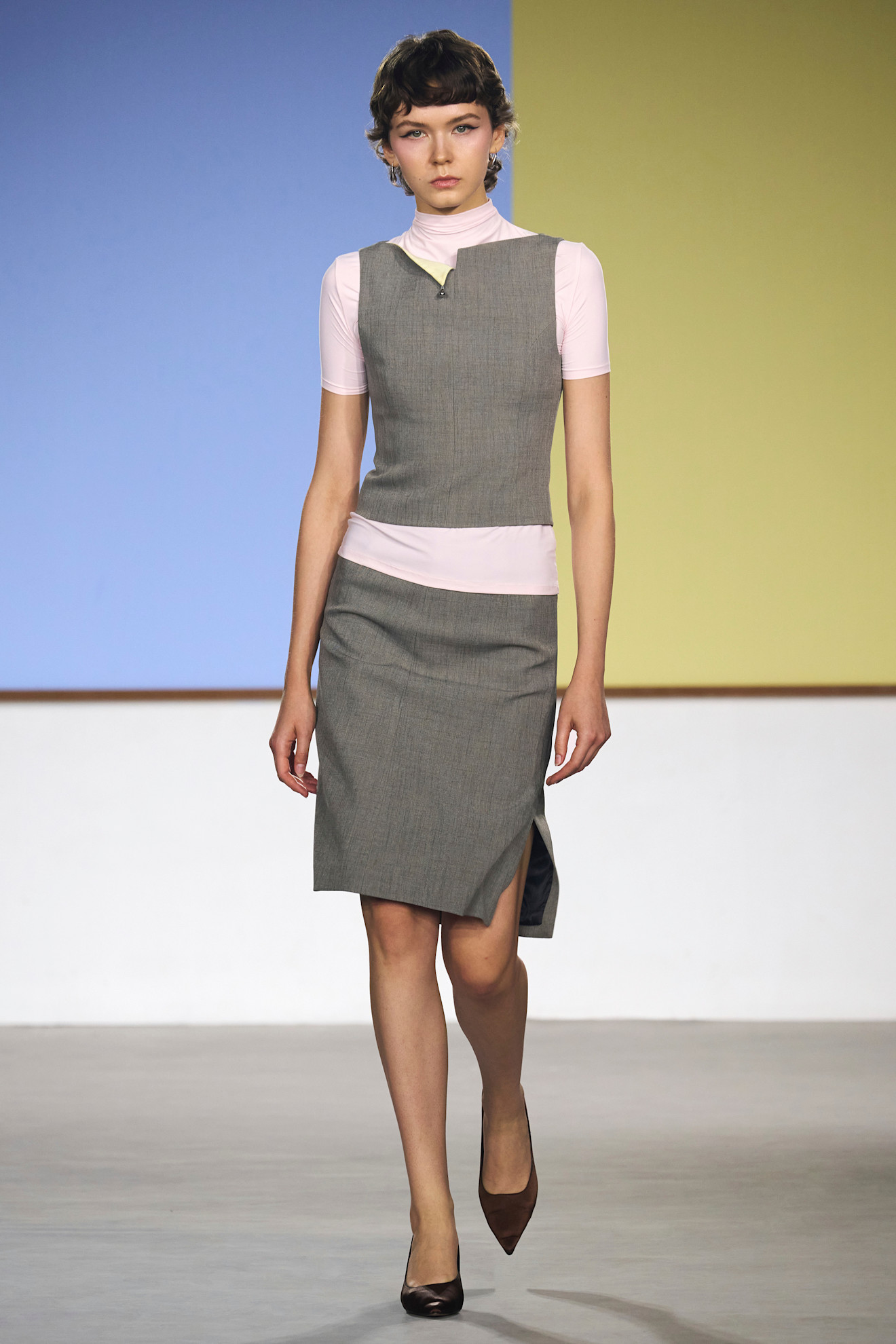 Layering staples for spring 2026 is shown in a photo from Yirantian's runway show, with a model walking while wearing a pink cap sleeve turtleneck sweater styled underneath a gray boatneck blouse styled with a gray pencil skirt and black pointed pumps 
