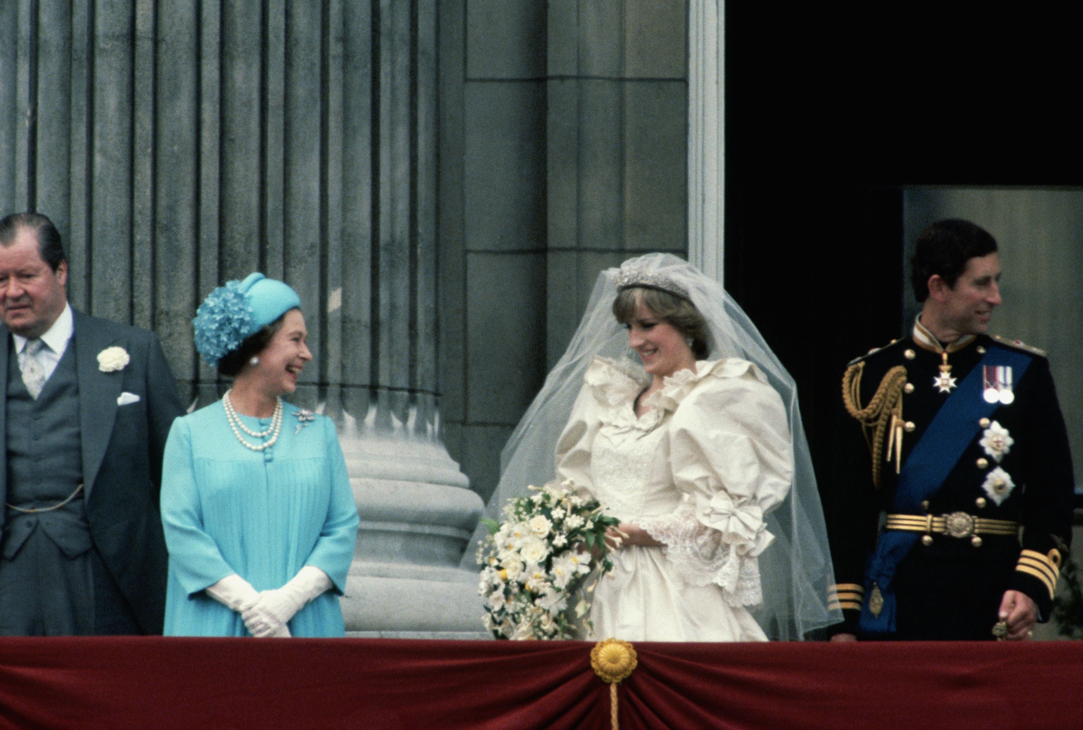 Queen Elizabeth wearing a blue coat and hat smiling at Princess Diana, who is standing on the balcony of Buckingham Palace with Prince Charles on their wedding day