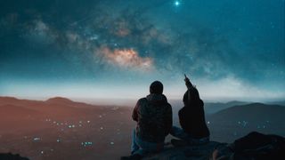 two people sit at a viewpoint overlooking a city in the distance and a starry sky above. the person on the right is pointing up to the sky.