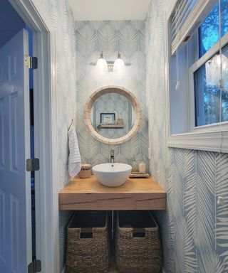 A bathroom with a wooden shelf with a white circular sink with two dark brown woven storage baskets underneath, a cream circiular mirror, silver wall sconces, and light blue wallpaper with white leaf illustrations
