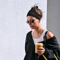 Hailey Bieber wears a black exercise outfit with sunglasses and a headband while carrying a cup of ice coffee. 