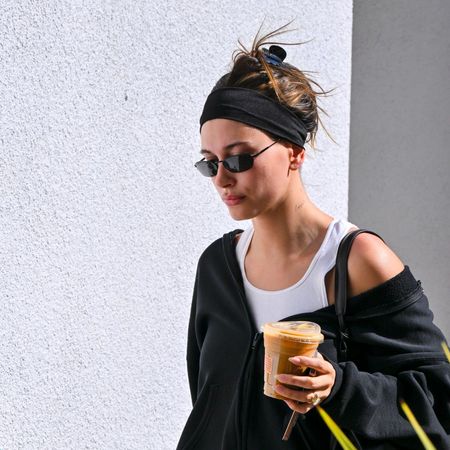 Hailey Bieber wears a black exercise outfit with sunglasses and a headband while carrying a cup of ice coffee. 