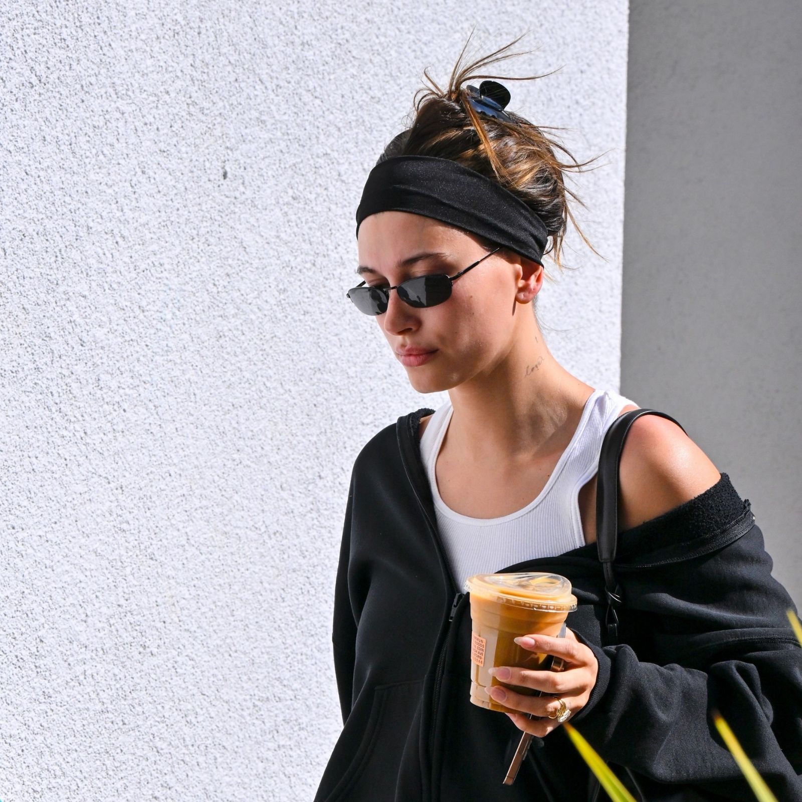 Hailey Bieber wears a black exercise outfit with sunglasses and a headband while carrying a cup of ice coffee. 