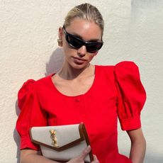 Elsa Hosk wears a red peplum top with black sporty sunglasses, gold earrings, and a beige Saint Laurent envelope clutch.