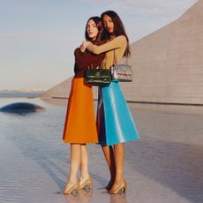 Two models stood in shallow water with their shoes submerged, hugging, wearing blue and orange skirts and Tory Burch handbags