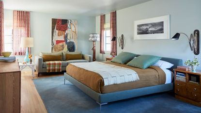 Image of a large, light blue bedroom with a bed in the middle and a sofa to the left. The bed is blue with a brown duvet and teal pillows. There are windows on the far wall that have short, red curtains.