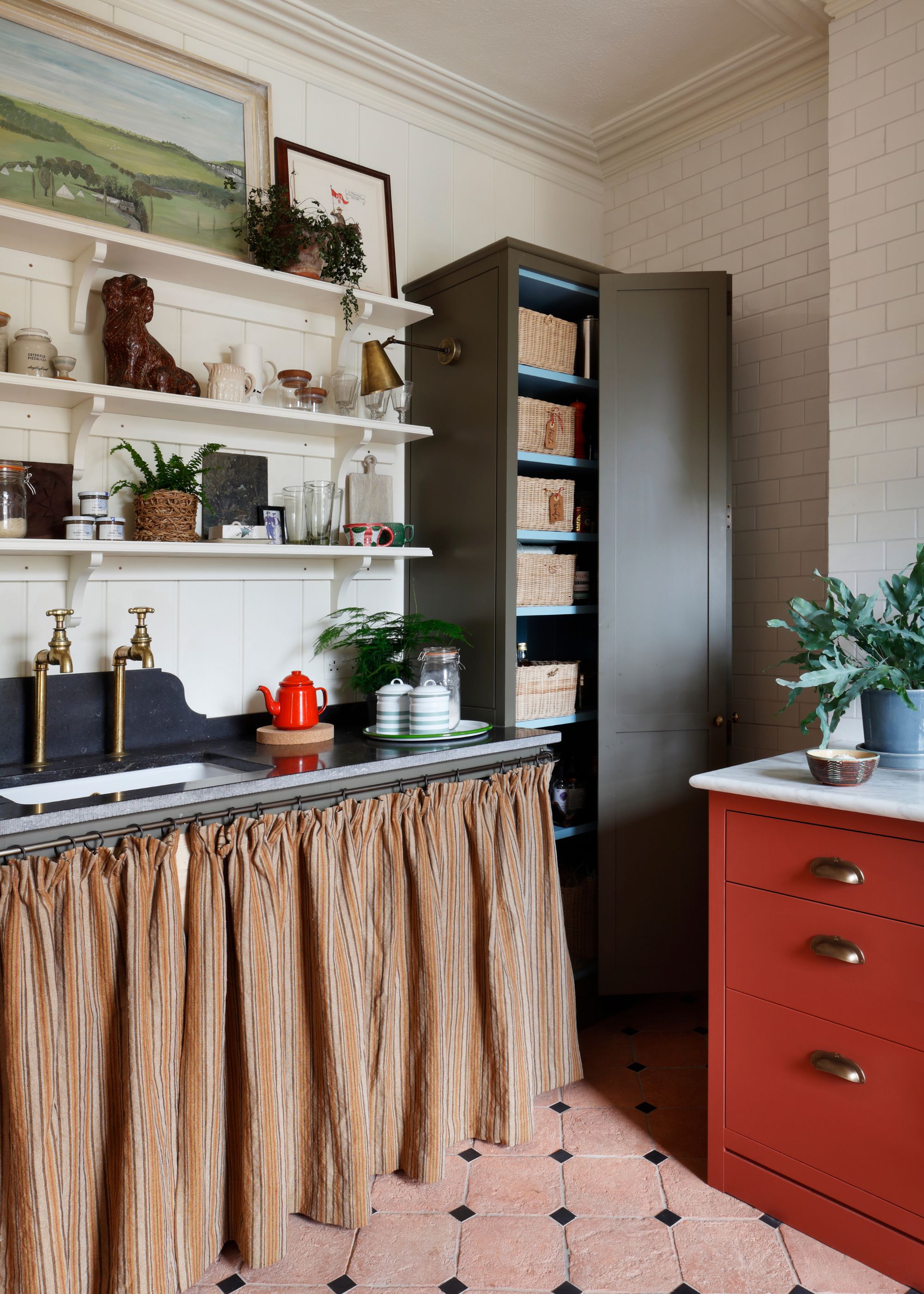 The corner of a rustic kitchen with terracotta tiled flooring, white subway tile walls, a sink with a linen cafe curtain beneath it, and a dark red cabinet. Open shelving above the sink and a freestanding, tall dark dresser in the corner.