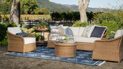 an outdoor patio with wicker outdoor furniture, white cushions, a blue outdoor rug, and a vineyard in the background