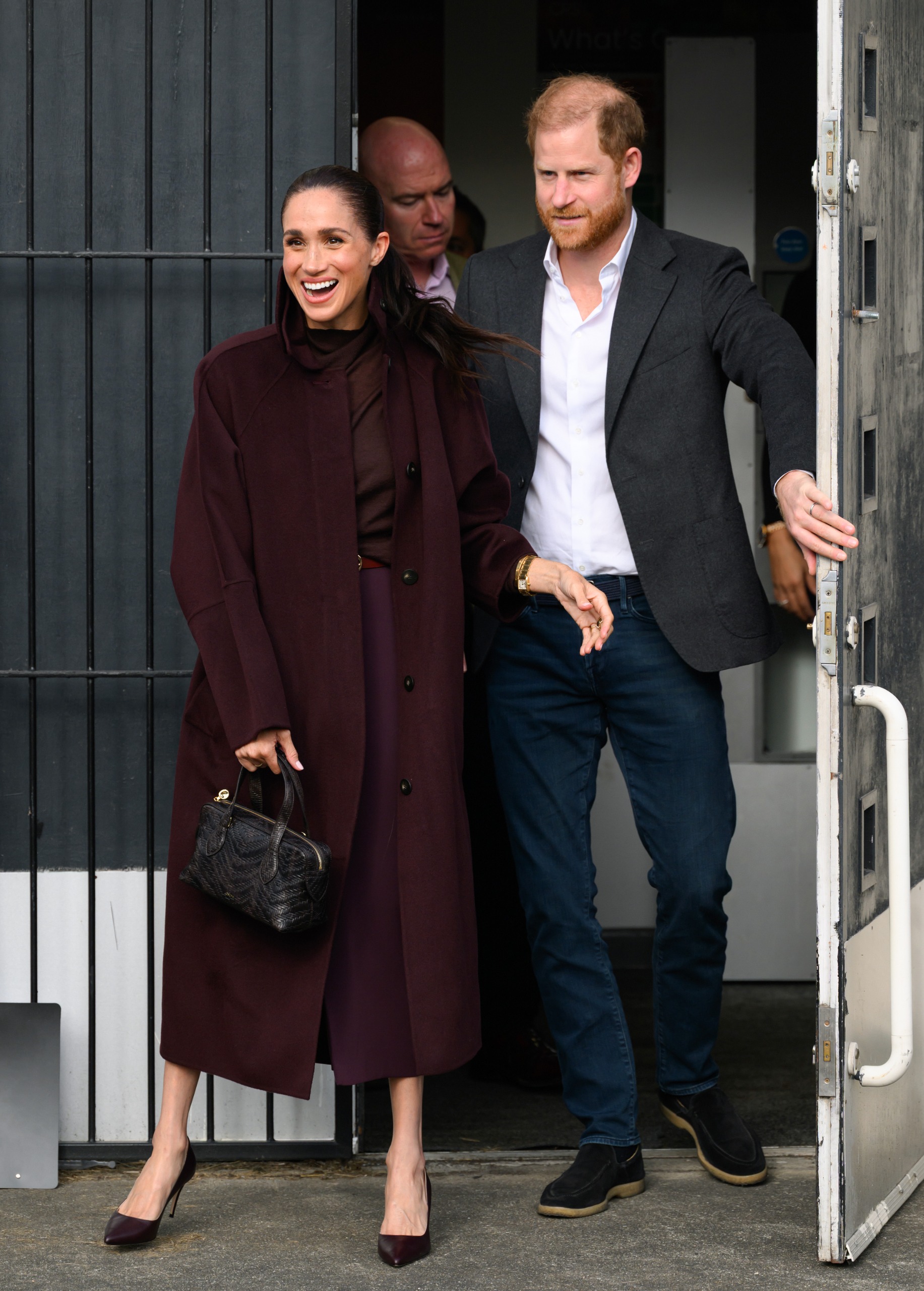 Meghan Markle wearing a marroon coat standing in front of Prince Harry