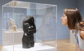 A small black sculpture of a naked woman's torso and head, displayed in a glass box. A woman is bending down to look more closely at the sculpture.