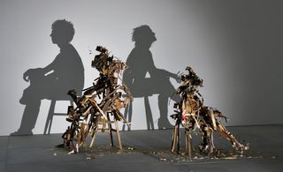 A closer view of one shadow sculpture made out of junk, metal, and wood. The light shines on the sculpture and casts a shadow on the wall. The shadow portrays a man and a woman sitting on stools with their backs turned to each other.