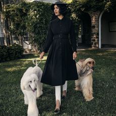 Nara wears black coat and white leggings while walking two luxurious-looking dogs on a leash in the front yard of a house