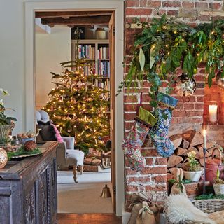 a brickwork fireplace with Christmas stockings and a lush garland hanging from it with a view into a room with a Christmas tree with twinkling fairy lights beside a traditional armchair