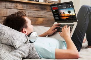 Teen boy with headphones works at laptop computer while lying in bed.