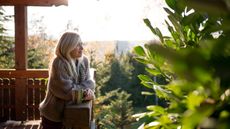 A smiling early retiree with a cup of coffee stands on her deck among a lush landscape.
