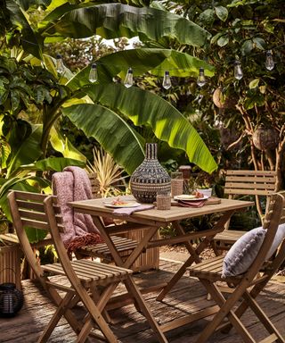 A small wooden outdoor patio dining table with chairs surrounded by palm plants in jungle luxe scheme
