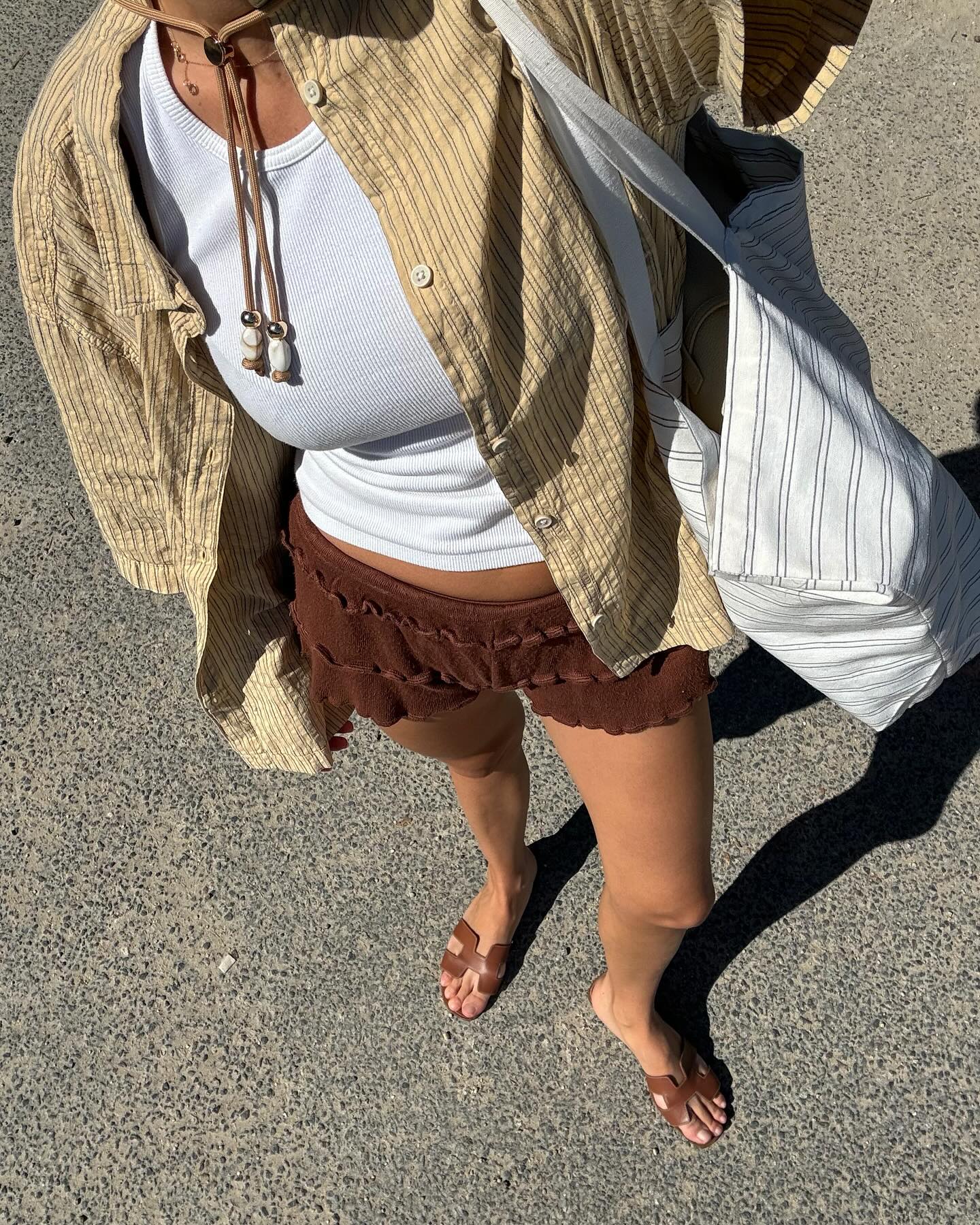 A woman taking an outfit selfie wearing a button-down shirt, a white tank top, brown short shorts, and brown Hermès flat slides.