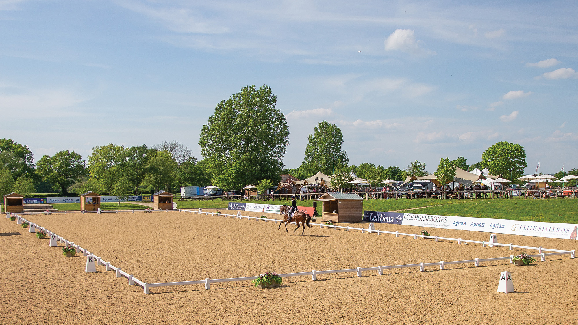 A distant shot of a dressage arena at Hickstead wth a horse cantering across the diagonal in front of the judges' huts.