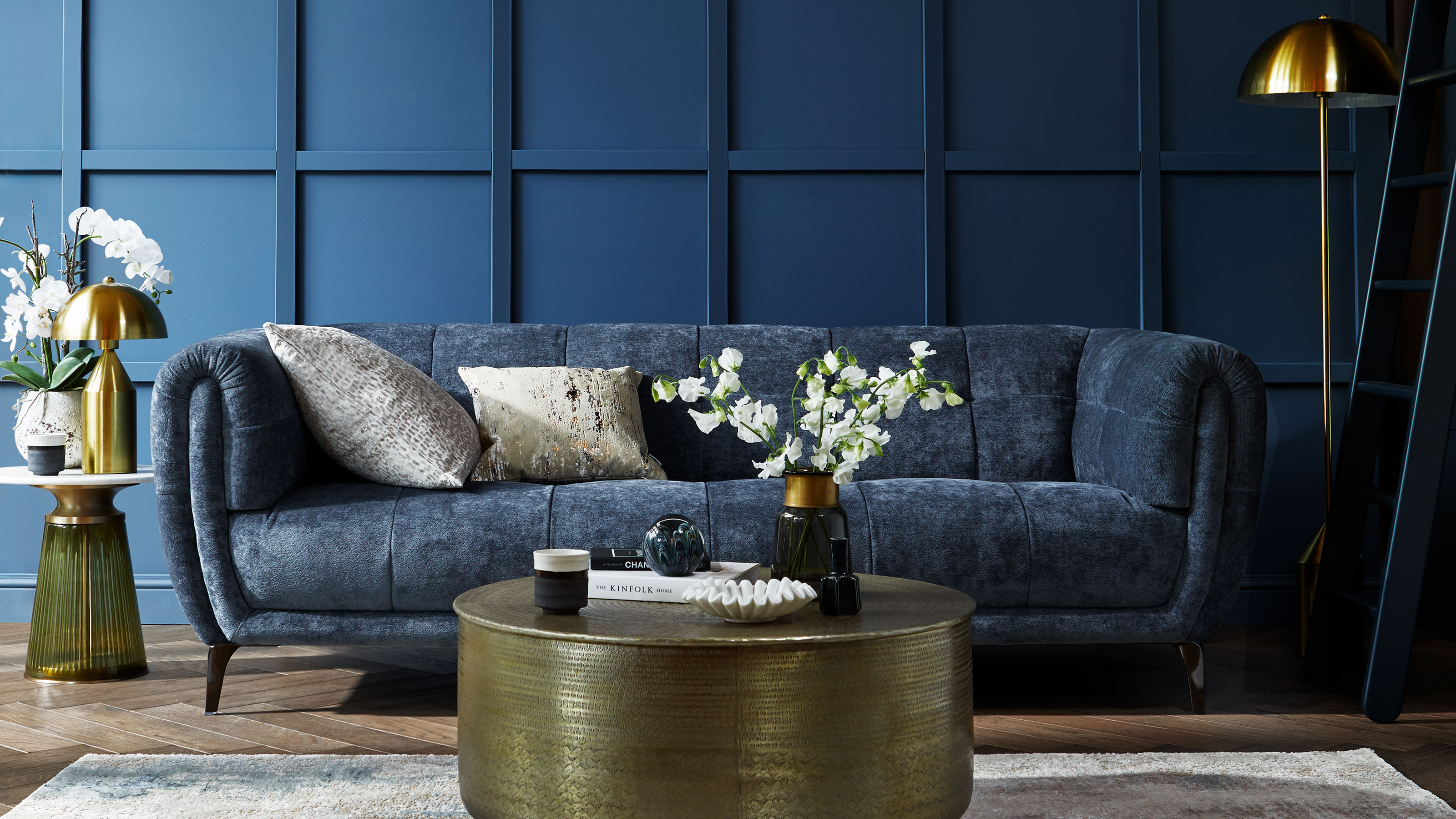 Blue sofa in front of a blue panelled wall, with a brass coffee table, gold floor lamp and gold table lamp