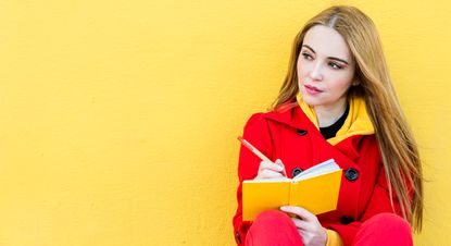Thoughtful woman wearing a red jacket sitting against a yellow wall while writing notes