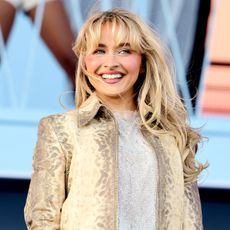 Sabrina Carpenter smiles and looks away from the camera wearing a beige jacket