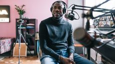 A man smiles while recording a podcast at his desk at home.