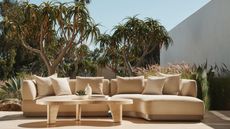 outdoor sofa styled with cushions with a travertine coffee table and large trees behind it