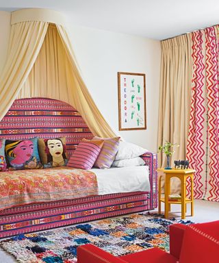 Tall pink patterned daybed with cream canopy, decorated with colorful cushions and throw, colorful checked rug, cream carpet, red armchair, pink patterned and cream curtains