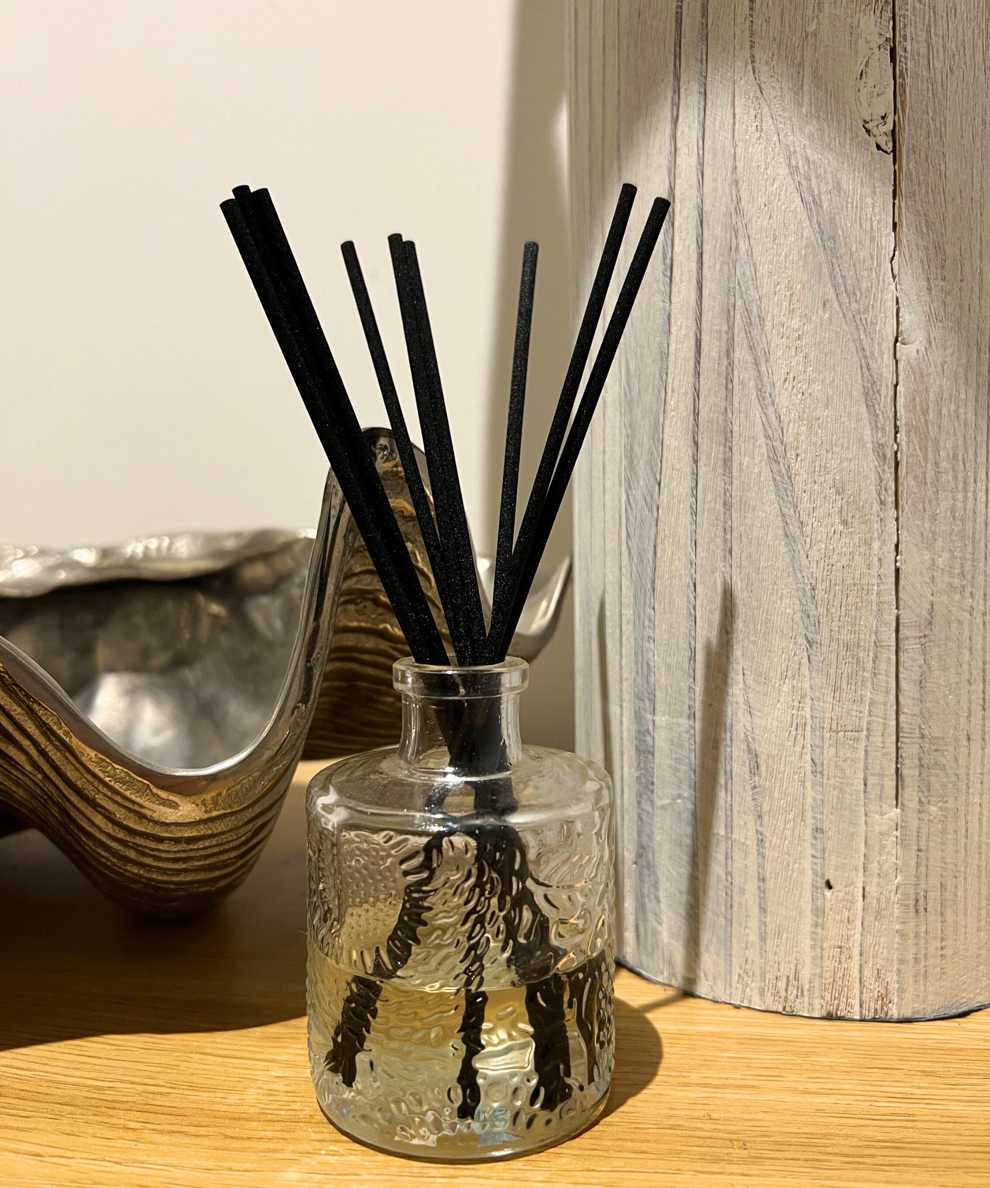 Voluspa diffuser on a light wood surface with a silver shell decoration behind and a white painted lamp, against a white wall.