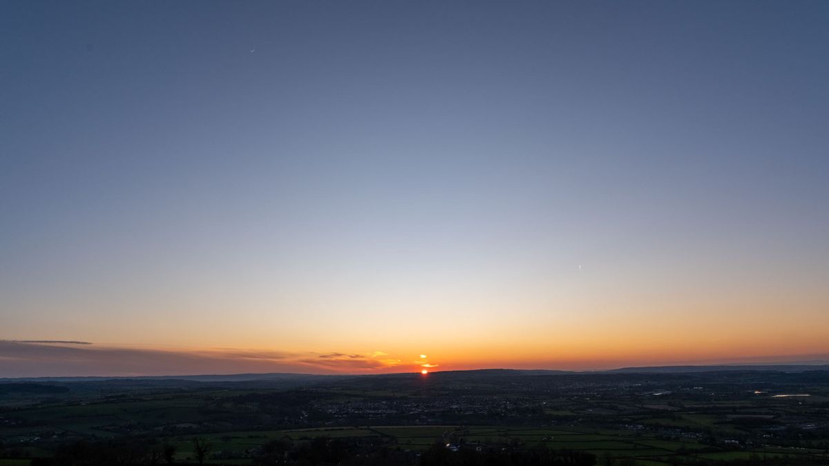 An image of a sunset over fields with an orange-and-blue sky and a crescent moon