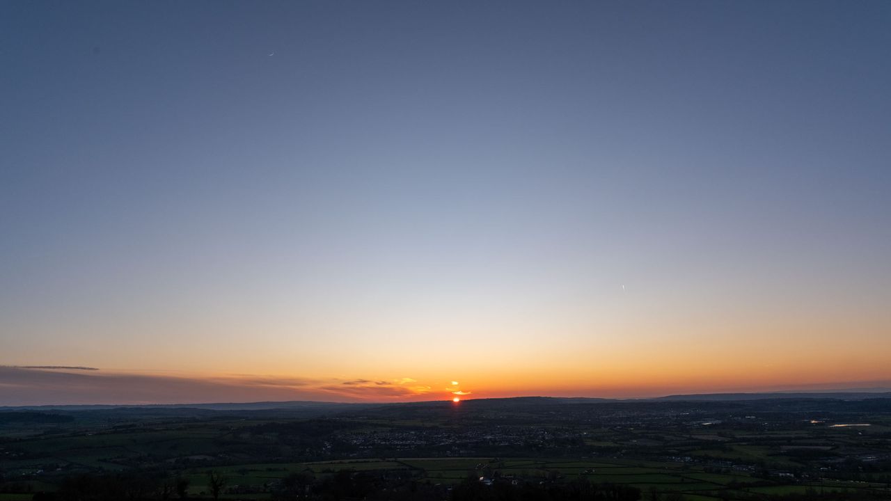 An image of a sunset over fields with an orange-and-blue sky and a crescent moon