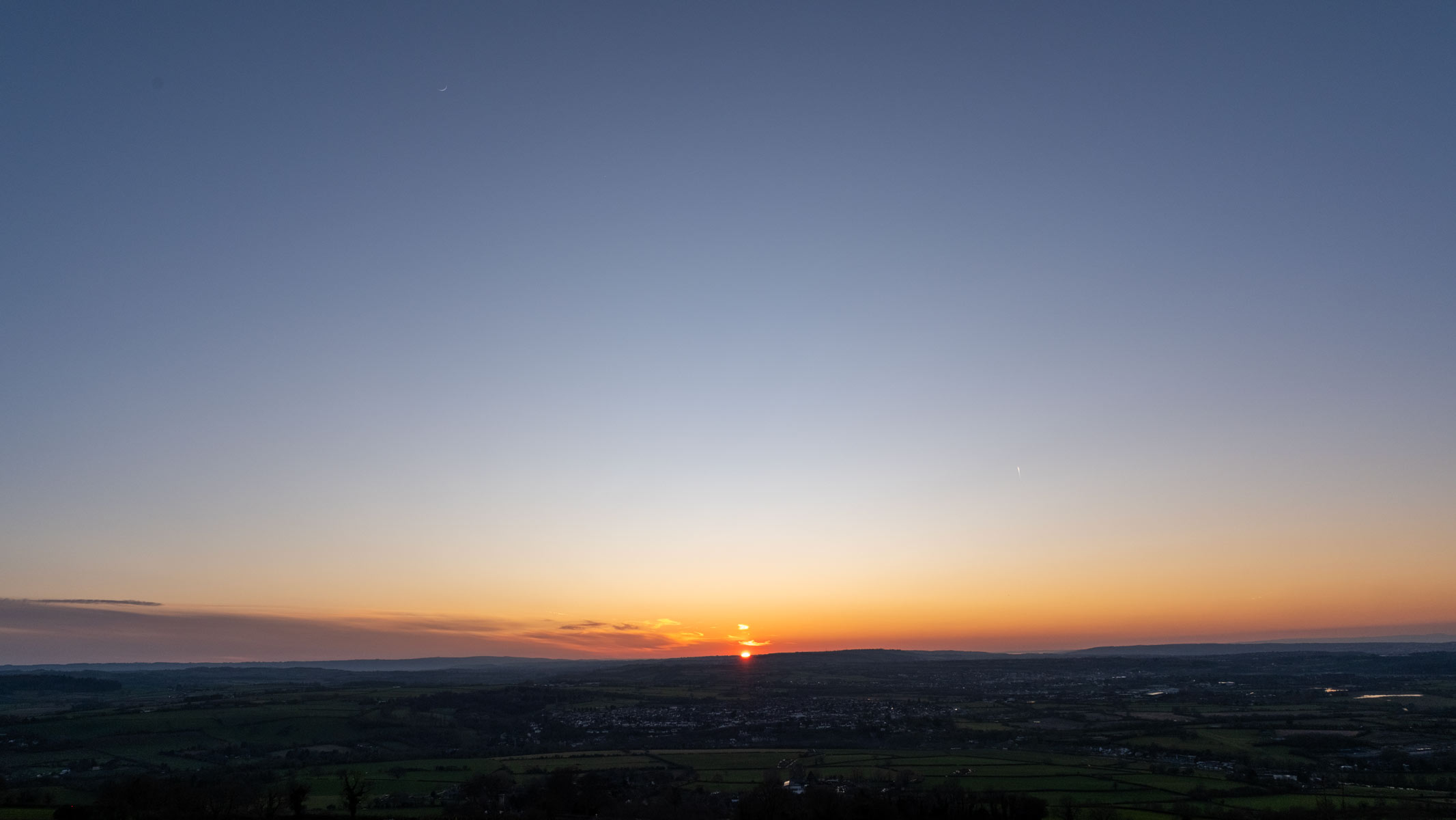 An image of a sunset over fields with an orange-and-blue sky and a crescent moon