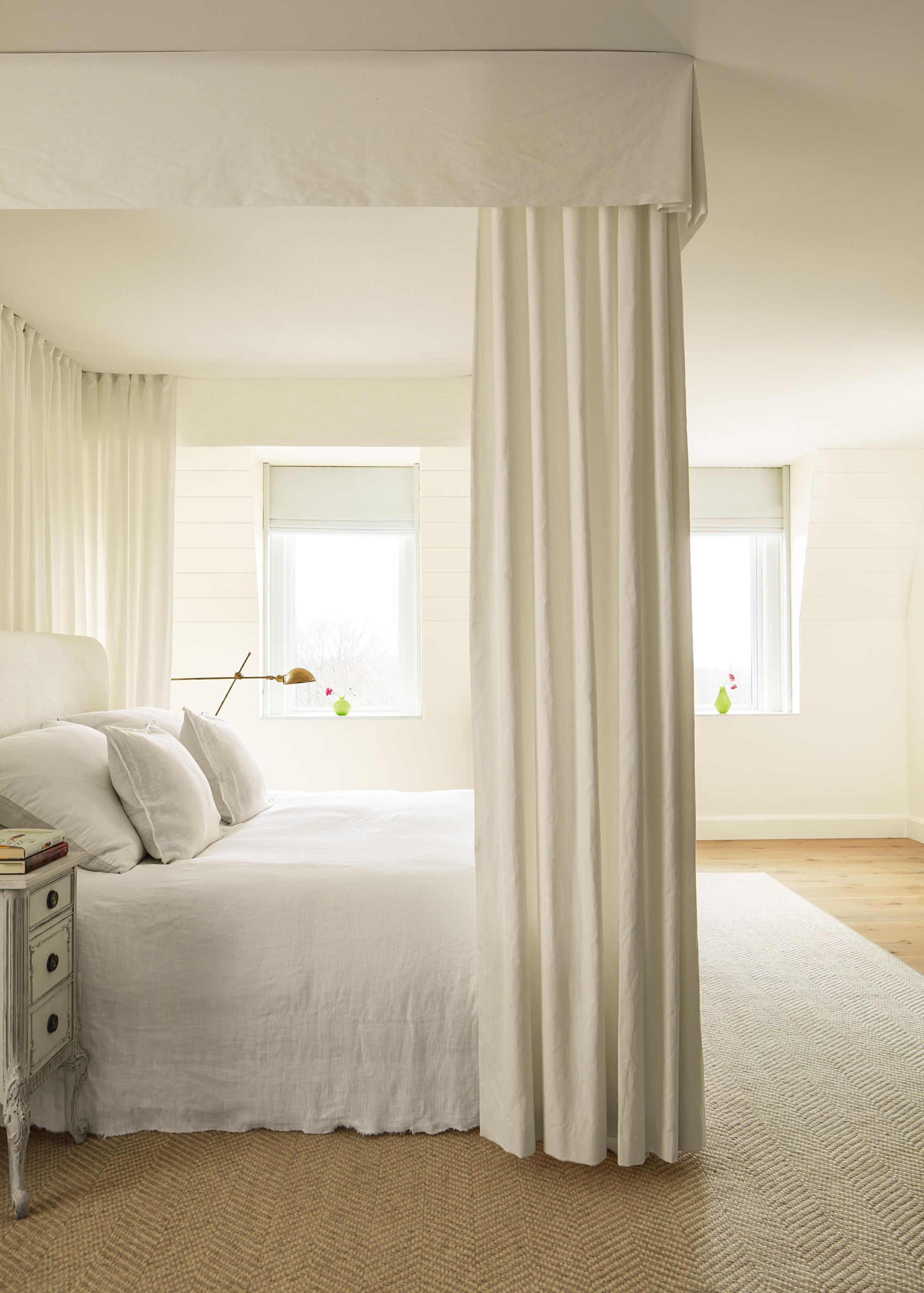 A calming all-white bedroom with canopy bed, neutral rug and white walls.