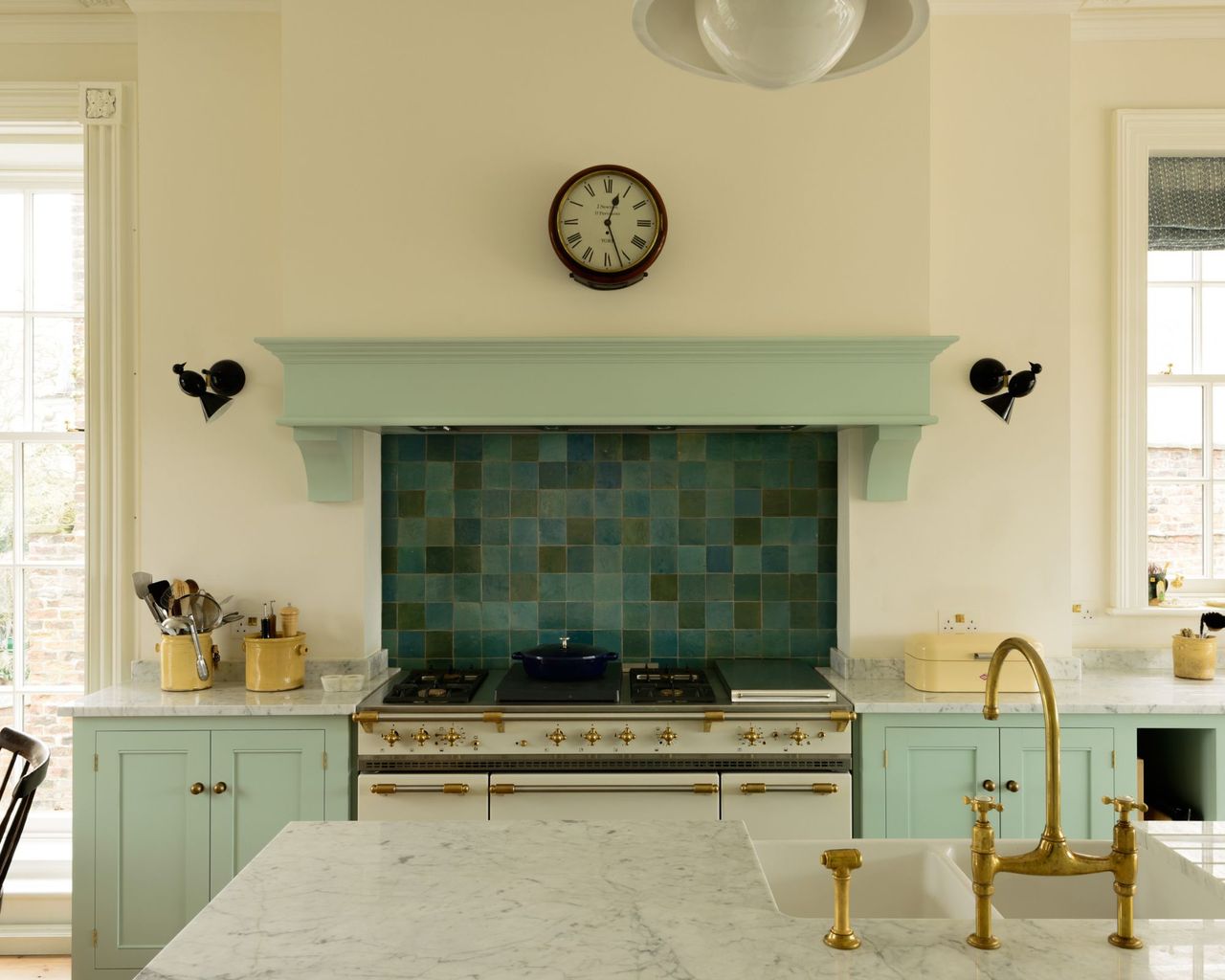 A bright white and duck egg blue kitchen with marble countertops, a gold tap, black light fixtures and large clock