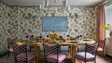 Breakfast room with botanical wallpaper, brass dining table, patterned dining chairs, blue chandelier and blush curtains.