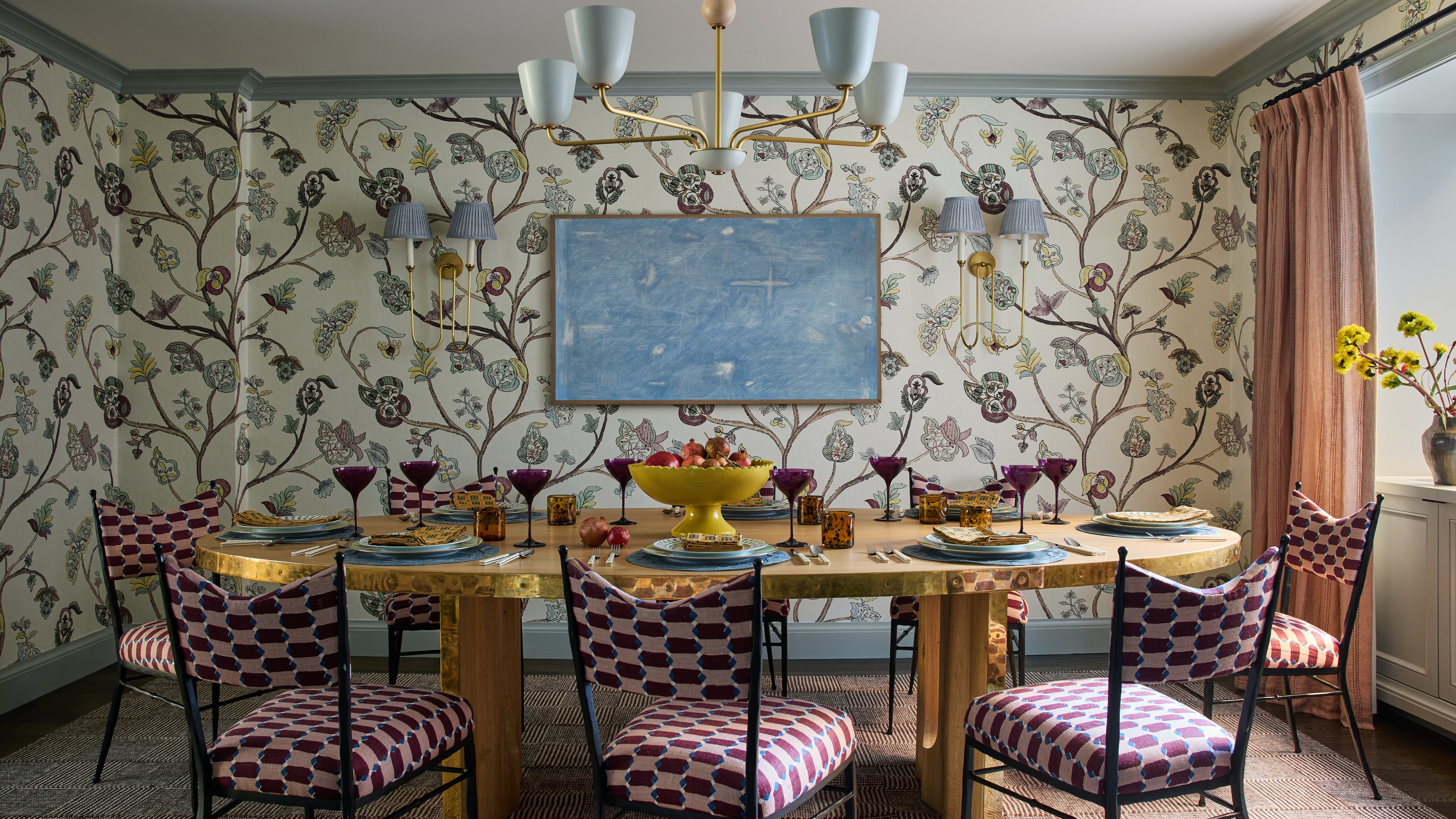 Breakfast room with botanical wallpaper, brass dining table, patterned dining chairs, blue chandelier and blush curtains.