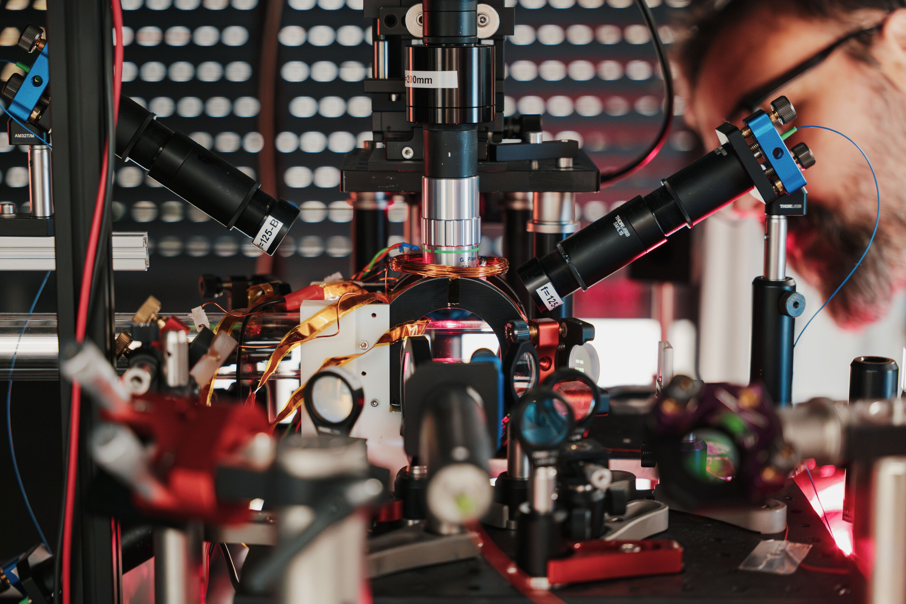 A man with glasses and a beard peers into an eye piece on a laser table with lenses and mirrors everywhere.
