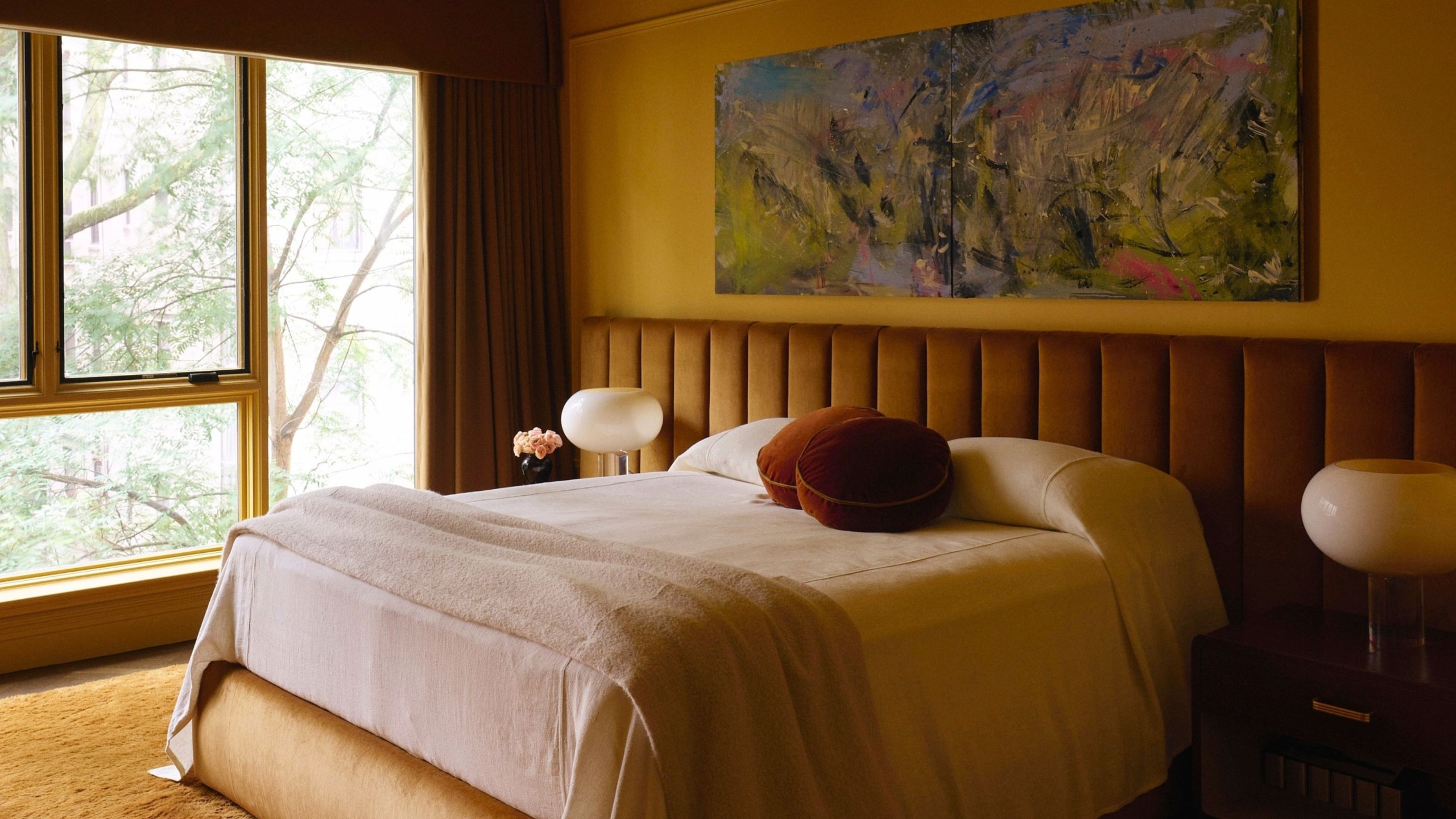 A modern bedroom with ochre color-drenched walls with decorative moldings, a large bed with neutral bedding, and a caramel-brown elongated headboard. A warm brown textured rug and large windows. 