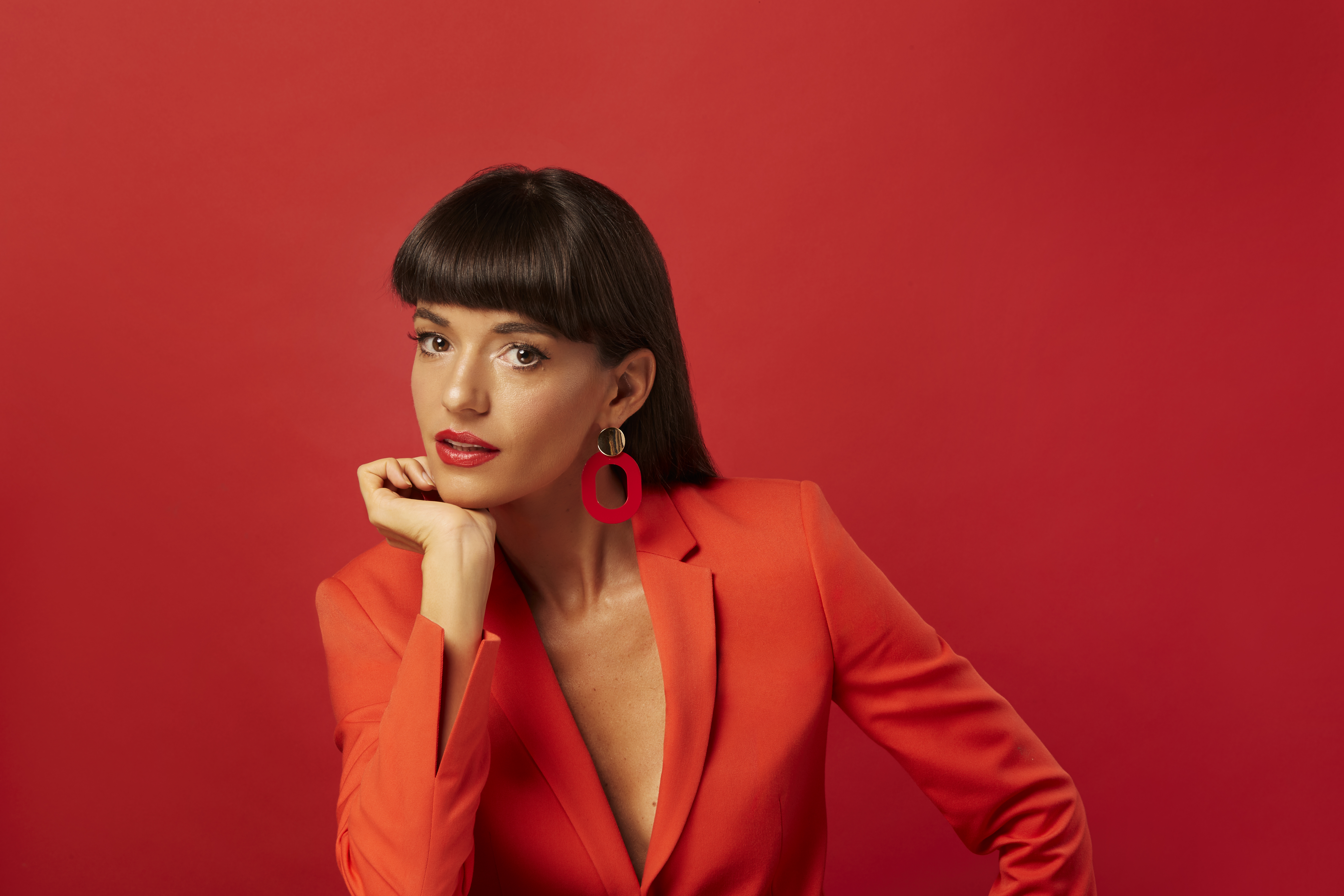 Jessica Walsh poses in a vibrant red blazer and matching large geometric earrings against a solid red background.
