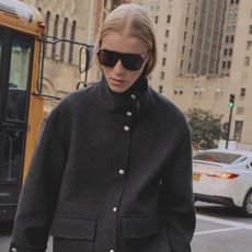 A Mango model wearing a gray funnel-neck jacket with silver buttons and large aviator sunglasses on the street in New York City.