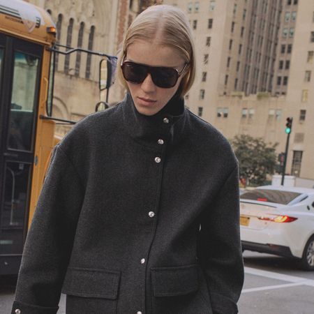 A Mango model wearing a gray funnel-neck jacket with silver buttons and large aviator sunglasses on the street in New York City.