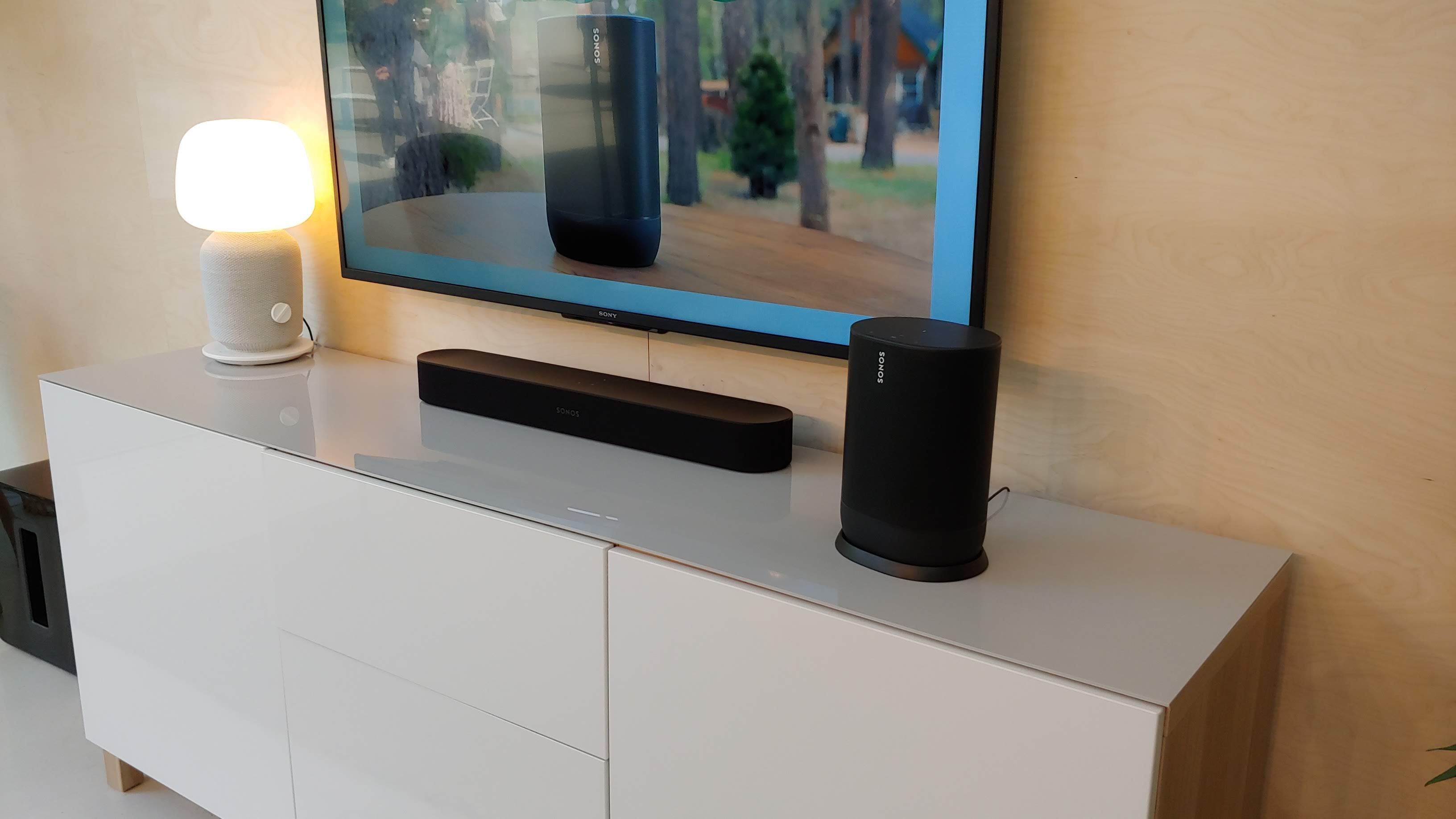 En svart Sonos Move står placerad på en vit TV-bänk bredvid en soundbar och en TV.
