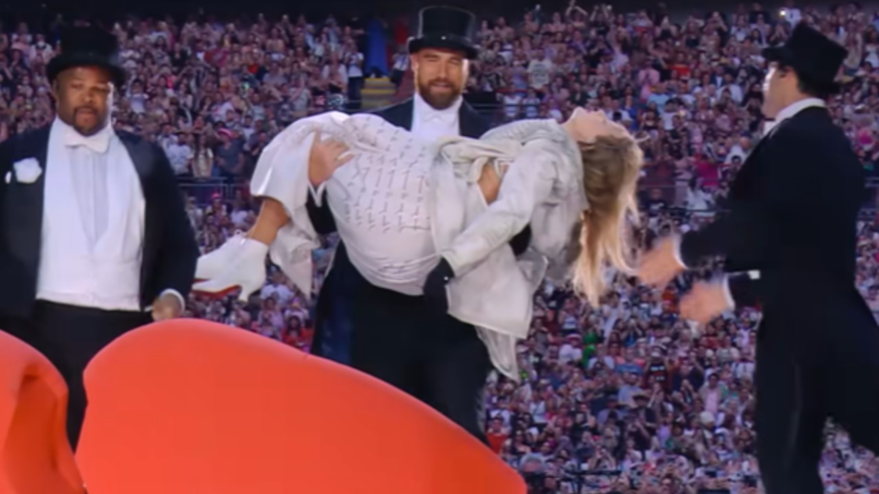 Travis Kelce carrying Taylor Swift to a red couch during the Eras Tour.