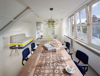 A room in the Hôtel Droog. To the right, large windows cover the entire wall. A long, dining table is set in front of them, with office-type chairs in navy blue. To the left is a sofa in grey, yellow and blue.
